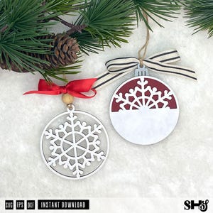 May include: Two circular Christmas ornaments featuring snowflake designs. One ornament is a white cutout snowflake with a red ribbon and wooden bead. The other is a solid white and maroon circle with a cutout snowflake top and a striped ribbon.