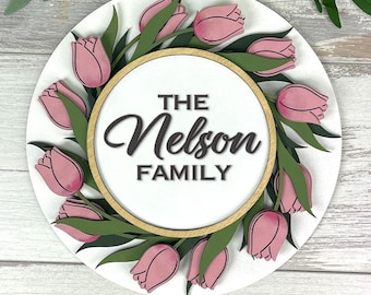 May include: A round, white wooden sign featuring a wreath of pink tulips and green leaves. The center circle has the text "THE Nelson FAMILY" in a dark brown script. The sign is a decorative item.