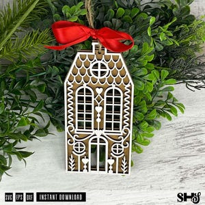 Puede incluir: Adorno navide&ntilde;o de madera con forma de casa, con intrincados detalles recortados. El adorno presenta una base marr&oacute;n con detalles blancos, una cinta roja y una cuerda de yute para colgar. Rodeado de follaje verde.
