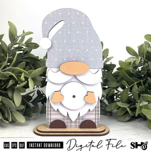 May include: A layered wooden gnome cutout featuring a light grey hat with white polka dots, a peach-coloured nose, a white beard, and a plaid patterned body. The gnome stands on a flat oval base and holds a circular white element with a small dot.