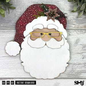 K&ouml;nnte beinhalten: Eine mehrschichtige Holzdekoration in Form eines Weihnachtsmann-Gesichts mit einer dunkelroten M&uuml;tze mit wei&szlig;em Sternenmuster, einem gro&szlig;en wei&szlig;en Bart, einem beigen Gesicht und einer gelben Brille. Eine glitzernde bronzene Schneeflocke ziert die M&uuml;tze.
