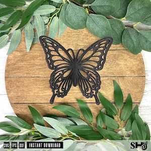 May include: A black butterfly cutout with intricate, symmetrical wing patterns featuring openwork details. The butterfly is positioned centrally on a round, light brown wooden surface with horizontal grain lines, surrounded by green leaves.
