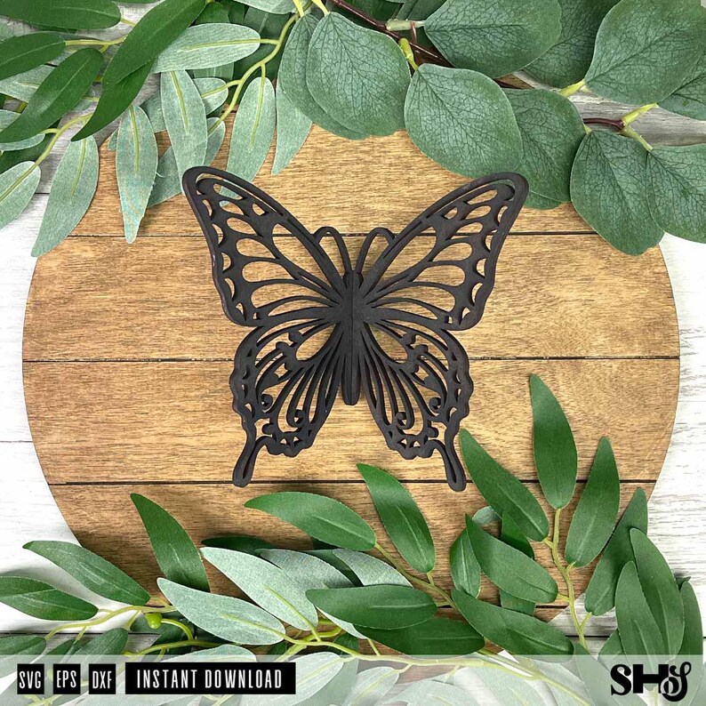 May include: A black butterfly cutout with intricate, symmetrical wing patterns featuring openwork details. The butterfly is positioned centrally on a round, light brown wooden surface with horizontal grain lines, surrounded by green leaves.