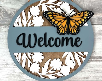 May include: A round wooden sign with a blue background and white floral cutouts. The sign says "Welcome" in black lettering and features a yellow butterfly with black markings.