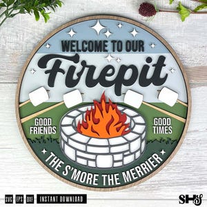May include: A circular layered wooden sign featuring a light blue sky with white stars, a green grassy field, and a stone firepit with orange and red flames. The text reads: WELCOME TO OUR Firepit, GOOD FRIENDS, GOOD TIMES, THE S'MORE THE MERRIER.