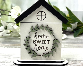 May include: A wooden house-shaped decoration with a black roof and a white body. The house features a green wreath around the words "home SWEET home." The base is white with a black trim. The image also includes the text "SVG EPS DXF INSTANT DOWNLOAD."