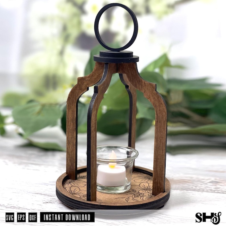 May include: A wooden lantern with a dark brown finish, featuring an arched frame, a circular top handle, and a base engraved with a floral pattern. A clear glass votive holder sits in the center, containing a small white LED tea light candle.