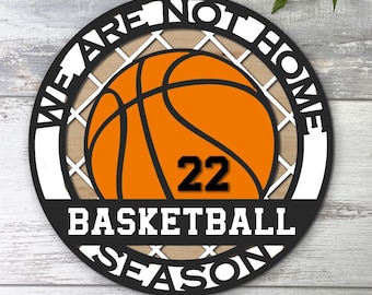 May include: Round wooden sign with the text "WE ARE NOT HOME BASKETBALL SEASON" in black and white. An orange basketball with the number 22 is in the center. The sign is hanging on a wooden background.