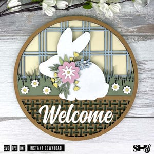 Pode incluir: Uma placa circular de madeira em camadas com a silhueta de um coelho branco adornado com uma flor rosa e folhas verdes. O fundo apresenta um padr&atilde;o xadrez azul e creme sobre uma &aacute;rea de relva verde com pequenas flores brancas e base tran&ccedil;ada.