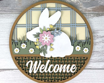 May include: A round, layered wooden Easter decoration featuring a white bunny with floral accents. The word "Welcome" is prominently displayed. The design includes a plaid background and grass, with a woven border.