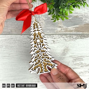 Puede incluir: Un adorno de &aacute;rbol de Navidad de madera con un contorno blanco y un intrincado dise&ntilde;o recortado. Est&aacute; adornado con un lazo de sat&eacute;n rojo y una percha de hilo. El adorno est&aacute; sostenido por las manos de una persona. La parte inferior de la imagen tiene el texto "SVG EPS DXF INSTANT DOWNLOAD".