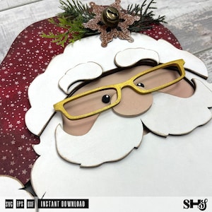 K&ouml;nnte beinhalten: Eine mehrschichtige Holzdekoration in Form eines Weihnachtsmanns mit roter M&uuml;tze mit wei&szlig;en Sternen, wei&szlig;em Bart und Schnurrbart sowie einer goldfarbenen Brille. Oben befindet sich eine glitzernde braune Schneeflocke mit kleiner Metallglocke und Tannennadeln.