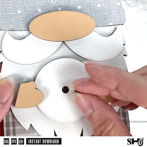 May include: A layered craft project featuring a gnome design with a grey patterned hat, a tan oval nose, a white beard, and a heart-shaped accent. The assembly includes a central white circular piece with a small hole and a metal fastener.