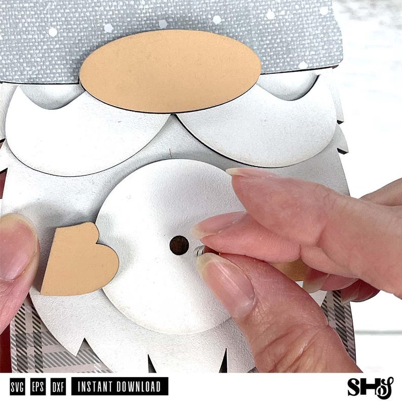 May include: A layered craft project featuring a gnome design with a grey patterned hat, a tan oval nose, a white beard, and a heart-shaped accent. The assembly includes a central white circular piece with a small hole and a metal fastener.