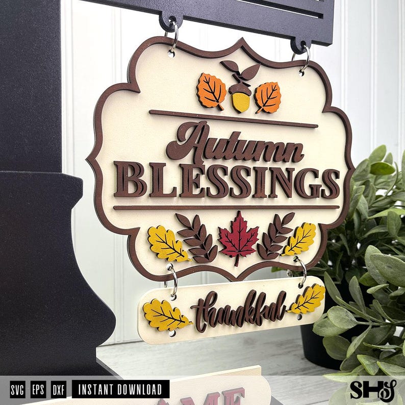 Op de afbeelding: Een decoratief hangend bord met de tekst Autumn BLESSINGS en thankful in bruine letters. Het bord is cr&egrave;mekleurig met gelaagde houten uitsparingen van oranje, gele en rode herfstbladeren en een eikel, opgehangen aan metalen ringen.