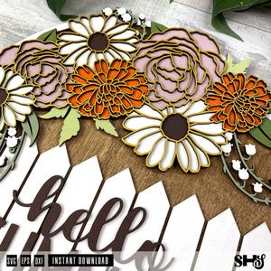 K&ouml;nnte beinhalten: Ein Holzschild mit einem wei&szlig;en Lattenzaun-Design und dem Wort "hello" in braunen Buchstaben. Das Schild ist mit einem Blumenarrangement aus wei&szlig;en, rosa und orangefarbenen Blumen verziert.
