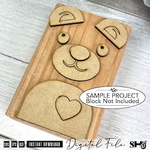 May include: A set of laser-cut wooden bear face components including two ears, two eyes, a nose with a mouth, and a belly piece with a heart cutout. The pieces are arranged on a rectangular wooden block. Text reads: SAMPLE PROJECT Block Not Included.