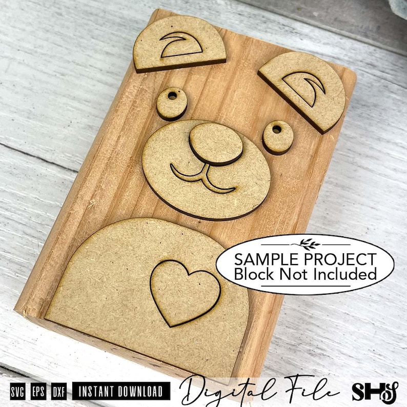 May include: A set of laser-cut wooden bear face components including two ears, two eyes, a nose with a mouth, and a belly piece with a heart cutout. The pieces are arranged on a rectangular wooden block. Text reads: SAMPLE PROJECT Block Not Included.
