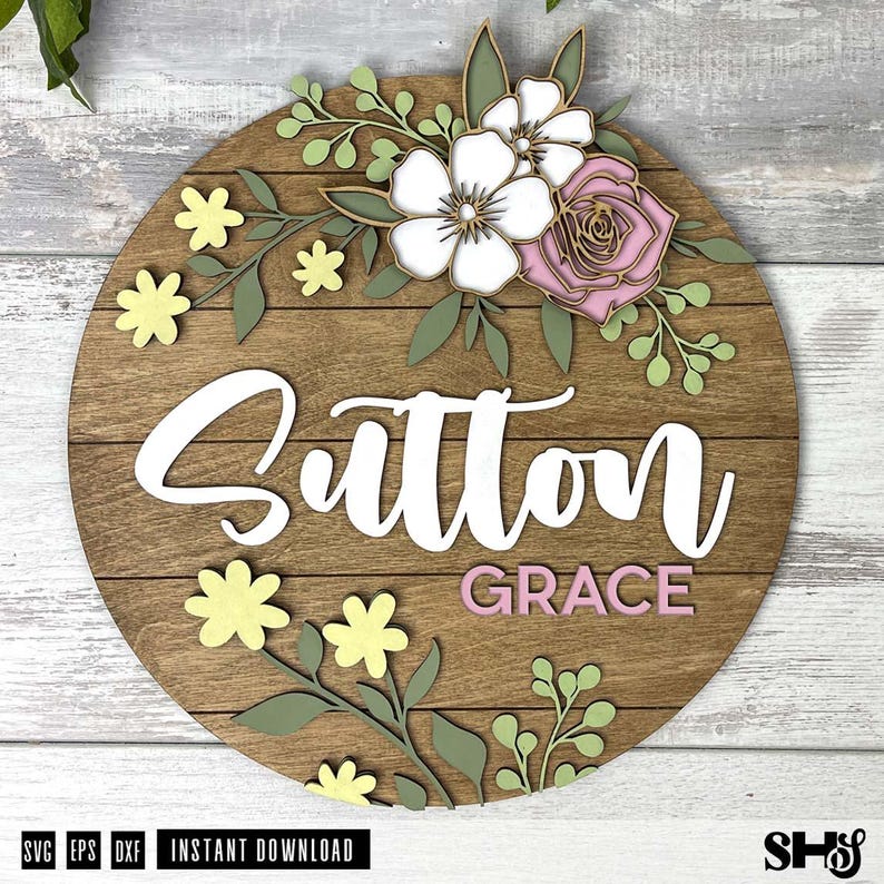 Puede incluir: Un letrero circular de madera con detalles de tablones horizontales y el nombre Sutton Grace en letras blancas y rosas. El dise&ntilde;o est&aacute; decorado con elementos florales de madera en capas en tonos blanco, rosa, amarillo p&aacute;lido y verde salvia.