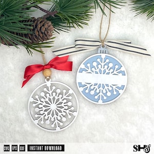 May include: Two circular snowflake ornaments. One is white with a red ribbon and wooden bead. The other has a light blue background, a white snowflake cutout, a horizontal white bar for personalisation, and a striped beige and black ribbon.