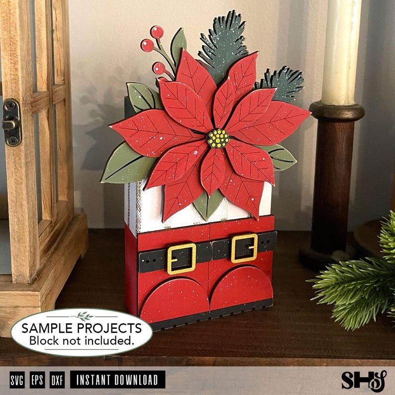 May include: A decorative wooden block display featuring a large red poinsettia flower with green leaves and berries at the top, sitting above a red base designed to look like Santa Claus boots with black belts and gold buckles.