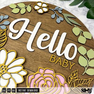 May include: A circular wooden sign with horizontal plank detailing featuring the white script text Hello and the gold block text BABY. The sign is decorated with layered cutouts of flowers and leaves in shades of sage green, gold, and soft pink.