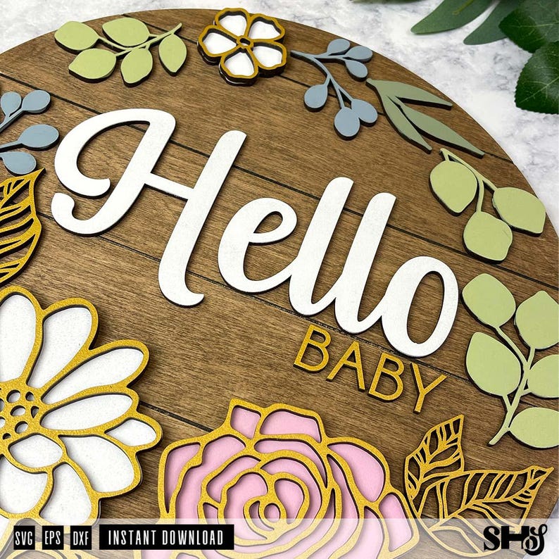 May include: A circular wooden sign with horizontal plank detailing featuring the white script text Hello and the gold block text BABY. The sign is decorated with layered cutouts of flowers and leaves in shades of sage green, gold, and soft pink.