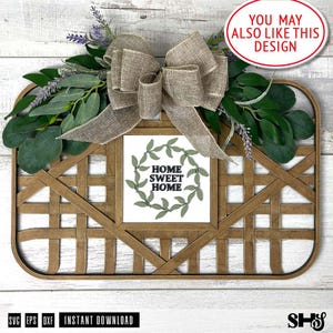 May include: A wooden sign with a woven frame and a burlap bow. The sign has a white background with a green wreath and the text "Home Sweet Home" in black.