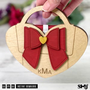 Könnte beinhalten: Ein herzförmiges Handtaschen-Ornament aus Holz mit einer großen, geschichteten roten Schleife und einem kleinen goldenen Herz in der Mitte. Die helle Holzoberfläche zeigt unten die eingravierten Buchstaben KMA und einen Griff.