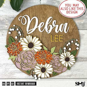 Puede incluir: Un letrero circular de madera con el nombre Debra en letra cursiva blanca y el texto LEE en letras de bloque amarillas. El letrero est&aacute; decorado con flores recortadas en capas, incluyendo margaritas blancas, crisantemos naranjas y peon&iacute;as.