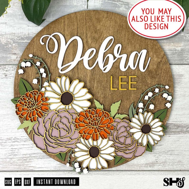 Puede incluir: Un letrero circular de madera con el nombre Debra en letra cursiva blanca y el texto LEE en letras de bloque amarillas. El letrero est&aacute; decorado con flores recortadas en capas, incluyendo margaritas blancas, crisantemos naranjas y peon&iacute;as.