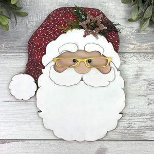 K&ouml;nnte beinhalten: Eine dekorative Weihnachtsmann-Gesichtsfigur mit einer dunkelroten M&uuml;tze mit wei&szlig;em Sternenmuster, einem gro&szlig;en wei&szlig;en Bart und Schnurrbart. Die Figur tr&auml;gt eine goldfarbene Brille und ist mit einer glitzernden braunen Schneeflocke verziert.
