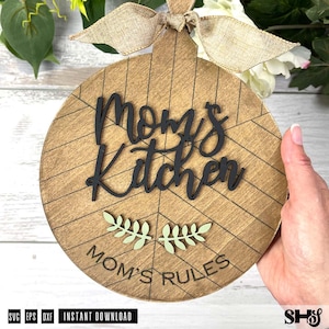 May include: A round wooden sign with a light brown finish and a geometric line pattern. It features the black 3D text "Mom's Kitchen" above two small light green leaf icons and the engraved text "MOM'S RULES" at the bottom, topped with a burlap bow.
