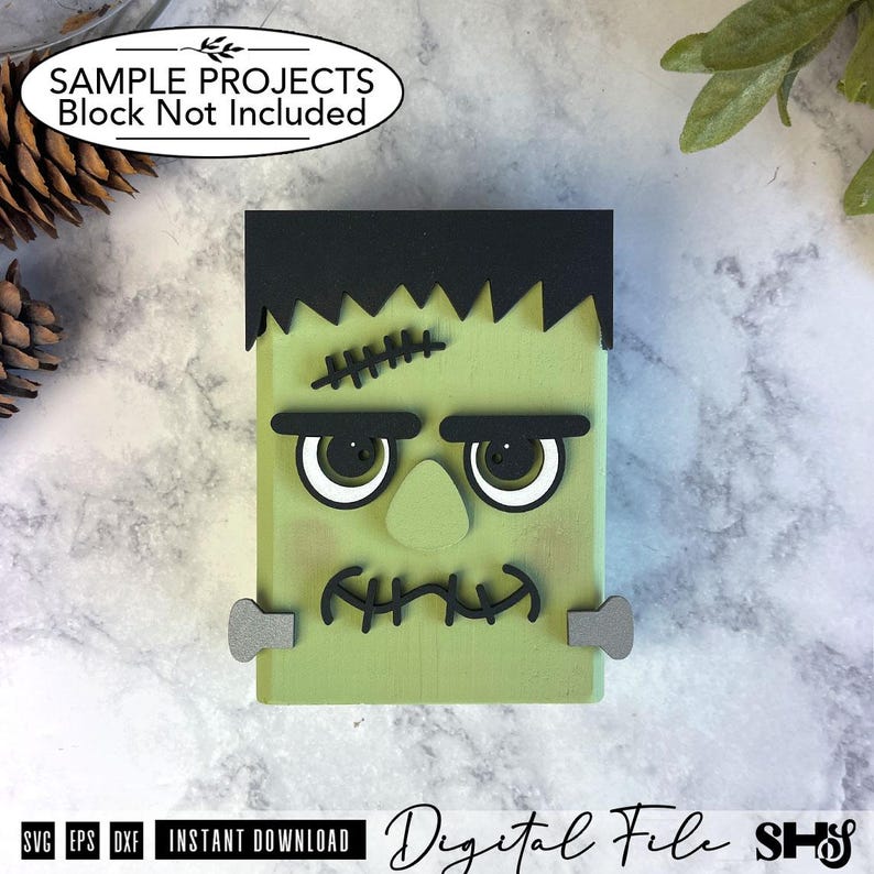 May include: A light green rectangular wooden block decorated as a monster face with black hair, thick black eyebrows, a central nose, and a stitched mouth. The design includes two grey bolts on the sides and a black stitched scar on the forehead.