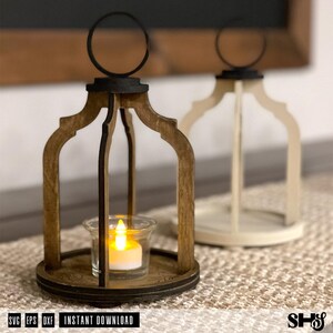 May include: Two decorative lantern frames with arched wooden supports and a circular top ring. One lantern is dark brown wood, the other is off-white. A clear glass votive holder with a glowing yellow LED tea light sits inside the brown lantern.