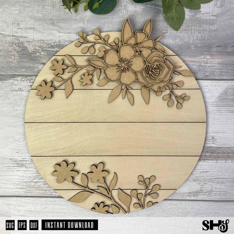Puede incluir: Un letrero circular de madera clara con l&iacute;neas de tablones horizontales y adornos florales cortados con l&aacute;ser. La secci&oacute;n superior muestra un grupo de flores y hojas, mientras que la secci&oacute;n inferior presenta un arreglo m&aacute;s peque&ntilde;o de flores.