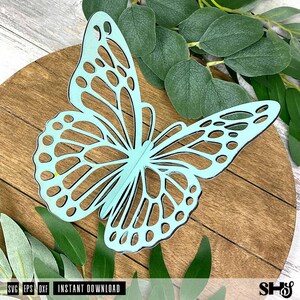 May include: A light blue, laser-cut butterfly silhouette featuring intricate, open-work wing patterns. The butterfly is displayed against a circular, dark brown wooden background with visible grain, surrounded by green eucalyptus leaves.