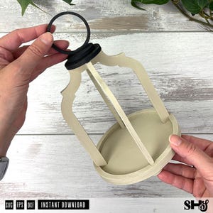 May include: A light beige wooden lantern frame with a circular base and a black ring handle at the top. The structure features four curved vertical supports connecting the base to the top cap, creating an open, cage-like design for a decorative piece.