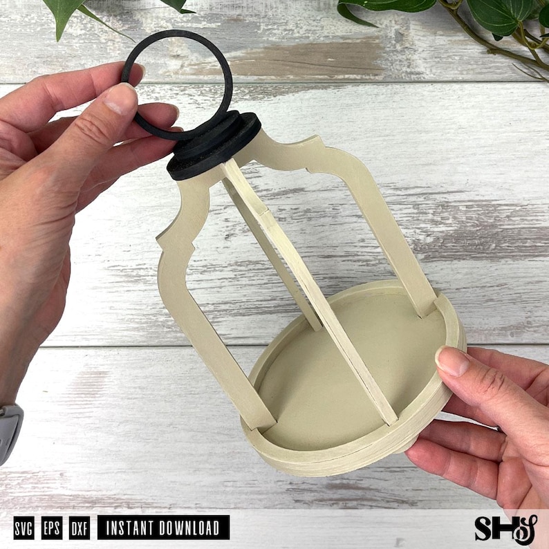 May include: A light beige wooden lantern frame with a circular base and a black ring handle at the top. The structure features four curved vertical supports connecting the base to the top cap, creating an open, cage-like design for a decorative piece.