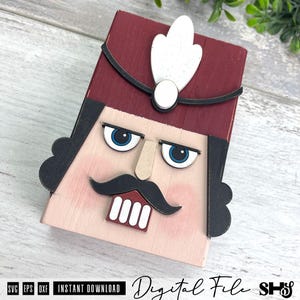 Puede incluir: Una decoraci&oacute;n de madera con forma de cara de cascanueces que presenta un sombrero rojo oscuro con una pluma blanca, cabello negro, ojos azules, nariz color canela, bigote negro y dientes blancos. El texto SVG EPS DXF aparece en la parte inferior.