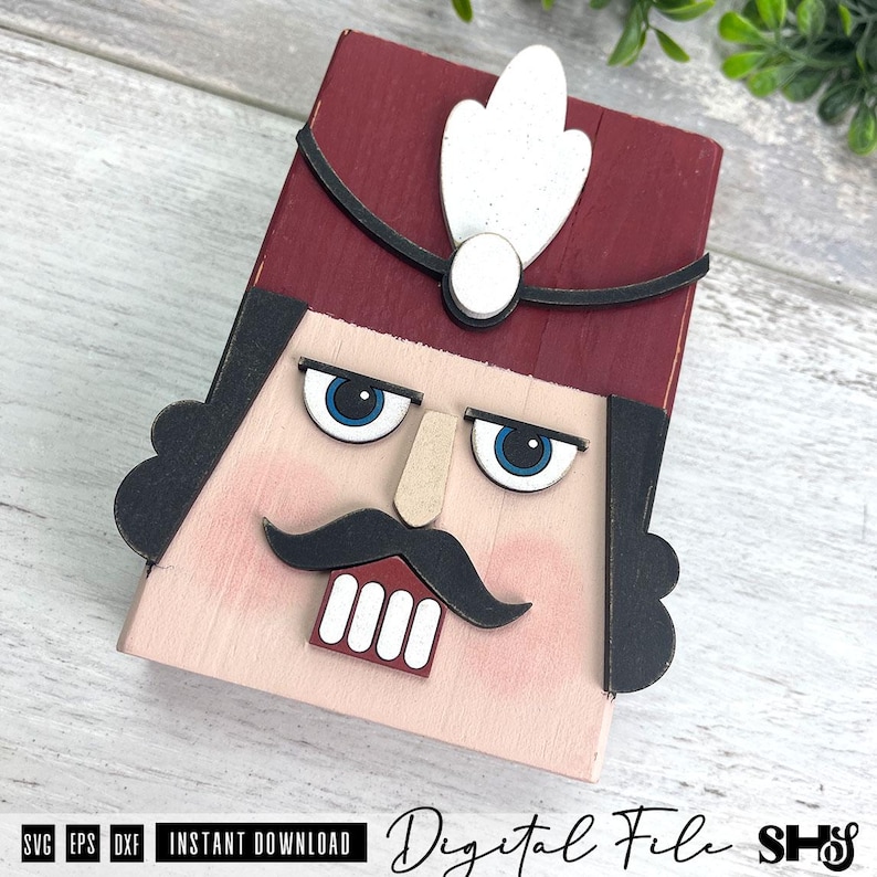 Puede incluir: Una decoraci&oacute;n de madera con forma de cara de cascanueces que presenta un sombrero rojo oscuro con una pluma blanca, cabello negro, ojos azules, nariz color canela, bigote negro y dientes blancos. El texto SVG EPS DXF aparece en la parte inferior.