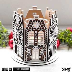 May include: A circular candle holder constructed from brown and white laser-cut panels that resemble a row of houses. The panels feature intricate white filigree patterns, arched windows, and heart motifs. A clear glass candle cup sits in the center.