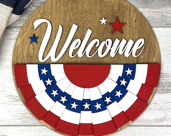 May include: A round wooden sign with the word "Welcome" in white script. The sign features a red, white, and blue bunting design at the bottom and stars. The sign is perfect for welcoming guests to your home.