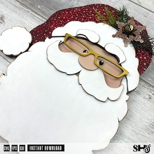 K&ouml;nnte beinhalten: Ein mehrschichtiger Holzausschnitt eines Weihnachtsmann-Gesichts mit einem gro&szlig;en wei&szlig;en Bart, einer roten M&uuml;tze mit wei&szlig;em Sternenmuster, einer goldfarbenen Brille und einem dekorativen braunen Glitzer-Schneeflocken-Akzent mit einer kleinen Metallglocke.