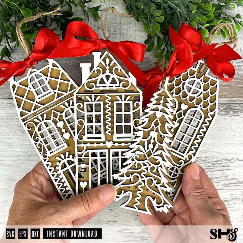 Puede incluir: Tres adornos navide&ntilde;os de madera cortados con l&aacute;ser con forma de casas de jengibre y un &aacute;rbol de Navidad. Cada adorno tiene un dise&ntilde;o superpuesto blanco, un respaldo marr&oacute;n y una cinta roja para colgar. Los adornos son sostenidos por manos.