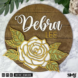 Op de afbeelding: Een houten bord met de naam "Debra Lee" in witte letters. Een witte roos met gouden contouren en groene bladeren is bovenop het bord gelegd.