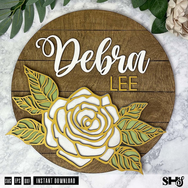Op de afbeelding: Een houten bord met de naam "Debra Lee" in witte letters. Een witte roos met gouden contouren en groene bladeren is bovenop het bord gelegd.