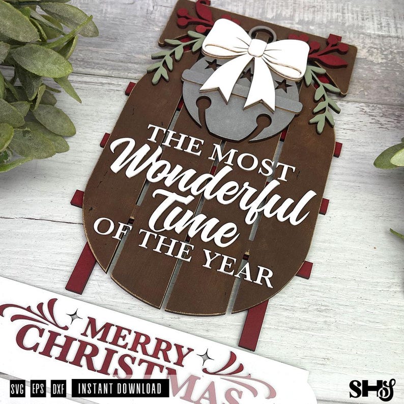 May include: A wooden sled-shaped wall sign featuring the white text THE MOST WONDERFUL TIME OF THE YEAR, a grey jingle bell with a white bow, and green leaf accents. A separate white banner below displays the red text MERRY CHRISTMAS.