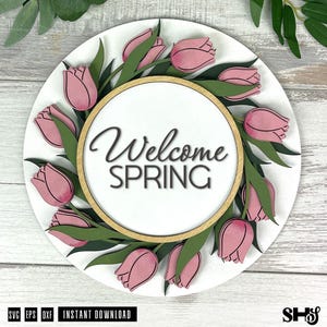 May include: A circular wooden sign featuring a white background with a wreath of layered pink tulips and green leaves. The center contains a light wood-toned circular frame surrounding the black text Welcome SPRING in a script and sans-serif font.