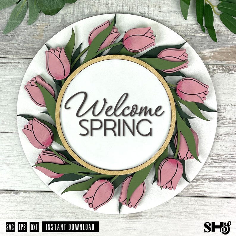 May include: A circular wooden sign featuring a white background with a wreath of layered pink tulips and green leaves. The center contains a light wood-toned circular frame surrounding the black text Welcome SPRING in a script and sans-serif font.